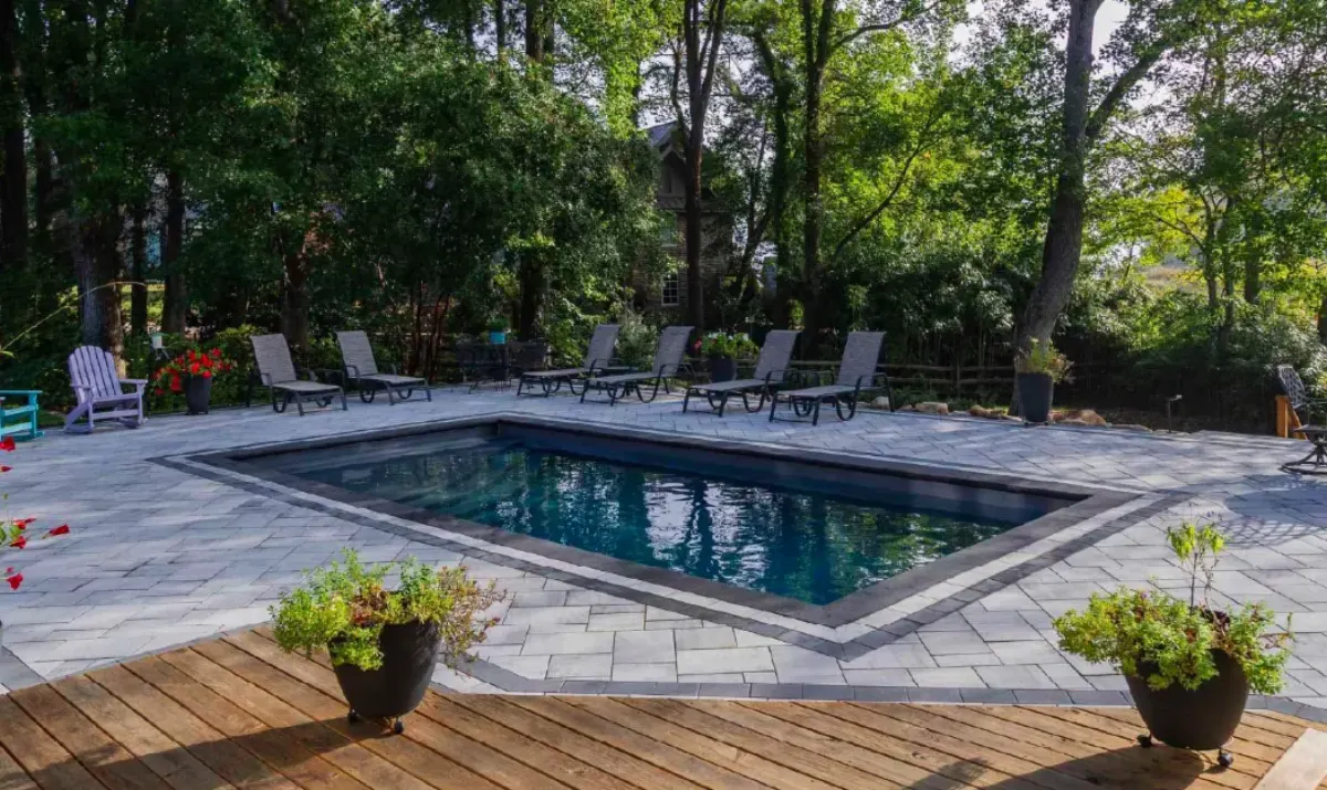 Inground fiberglass pool with large-format Unilock paver deck, wood platform and chaise lounges in a backyard surrounded by mature trees