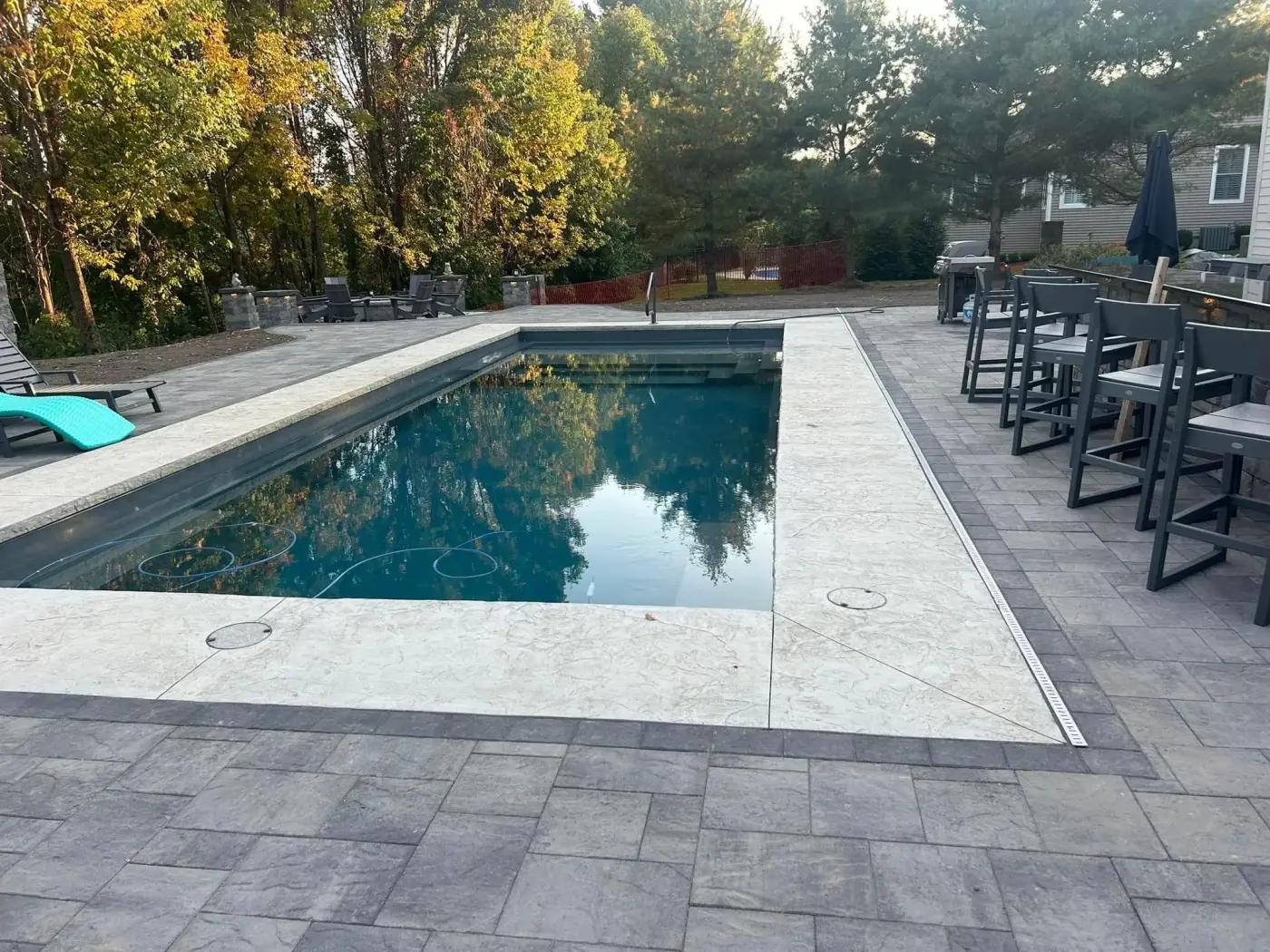 Rectangular inground pool with stamped concrete deck, paver patio, outdoor bar with stools and autumn trees in background
