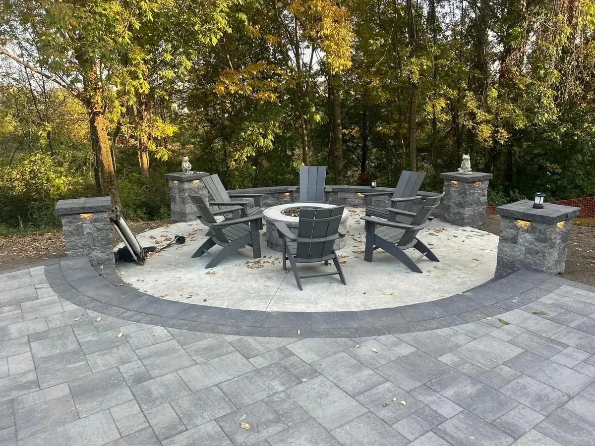 Circular paver fire pit area with Adirondack chairs, lit stone pillars and stone seating walls surrounded by fall foliage