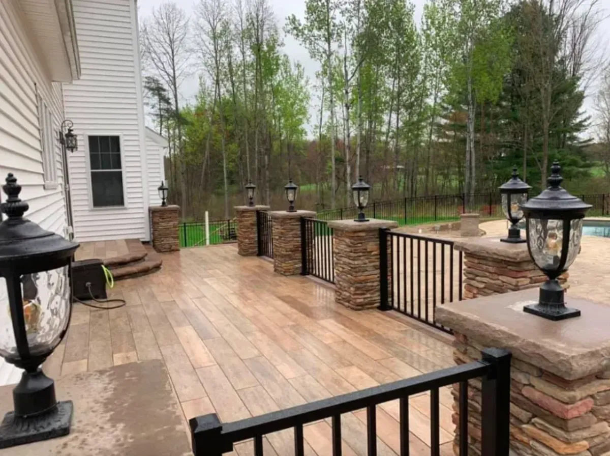 Wood-look porcelain tile patio with stacked stone pillars, lit lanterns and metal railing leading to a pool