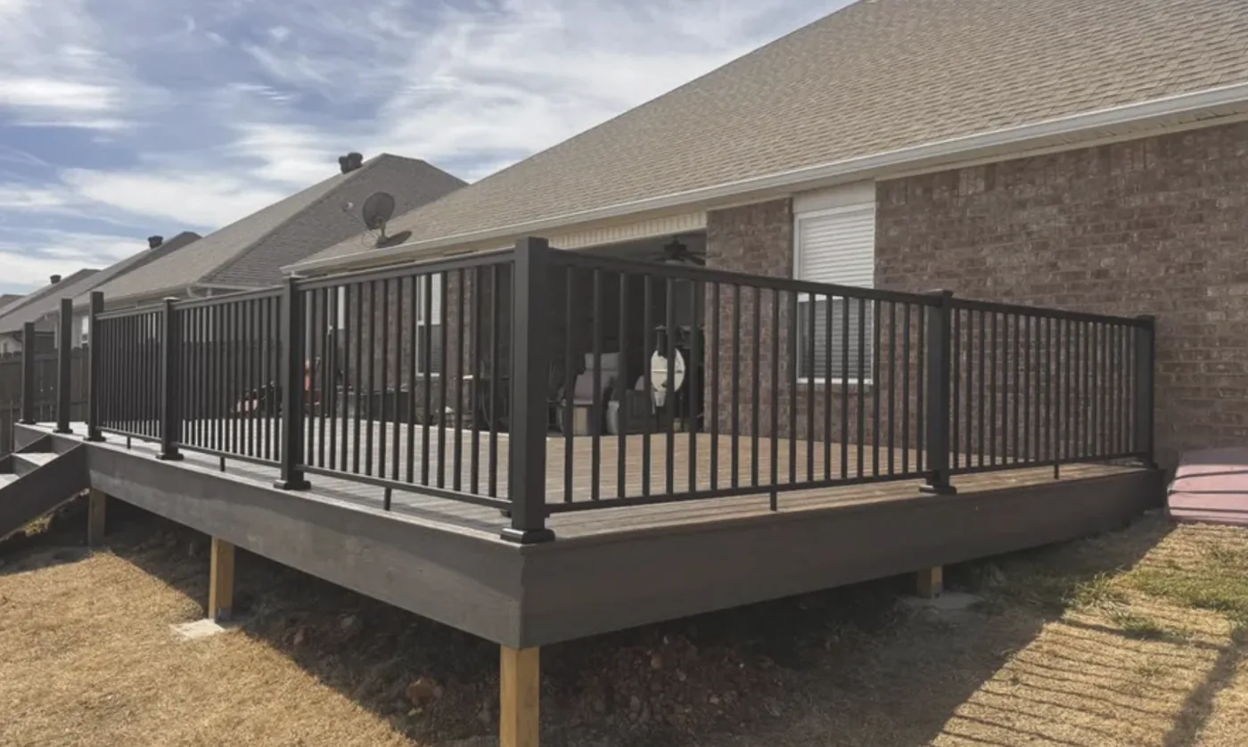 Composite deck with black aluminum railing attached to a brick home, raised on wood posts over a sloped backyard