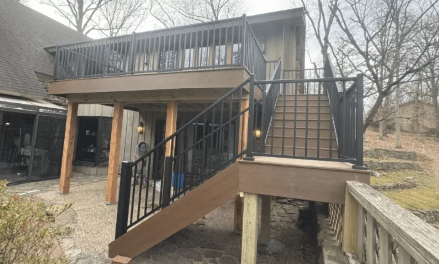 Two-story composite deck with staircase and black aluminum railing on a wooded hillside home